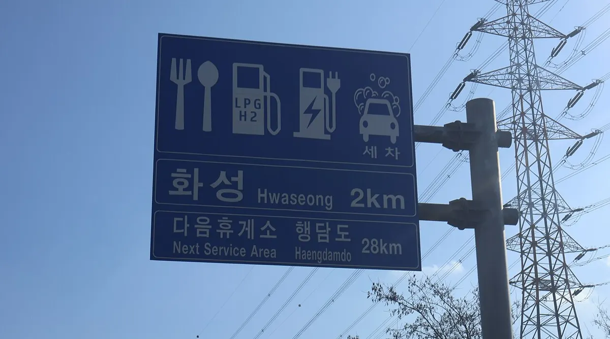 고속도로의 화성 휴게소 안내 표지판. 식당, 주유소, 전기차 충전, 세차 픽토그램과 함께 다음 휴게소까지의 거리가 표시되어 있습니다.