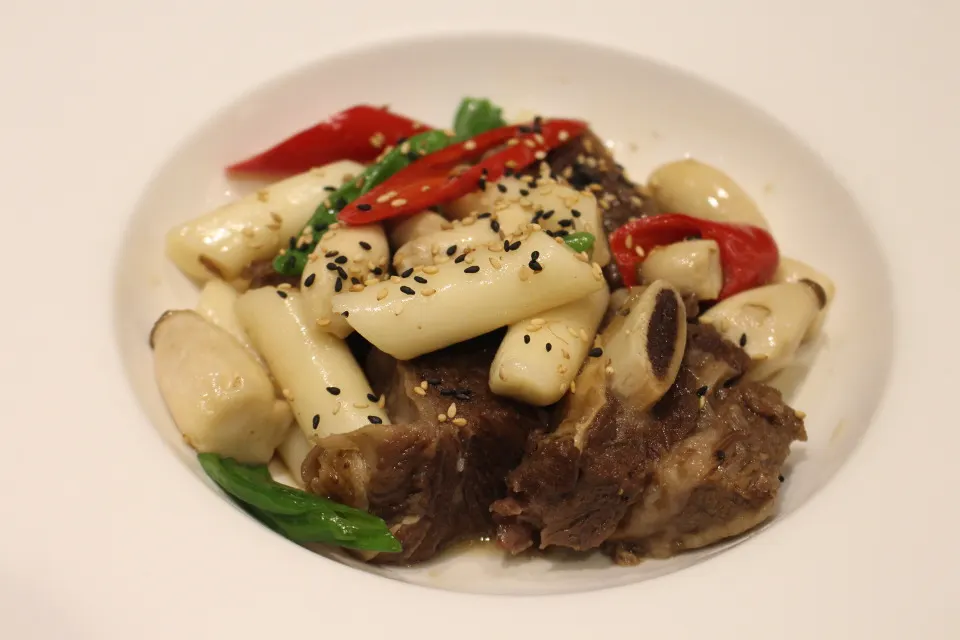 흰 접시에 담긴 소갈비찜. 부드럽게 익은 갈비 위에 가래떡 모양의 떡과 홍고추, 청고추, 통깨가 올라가 있는 모습