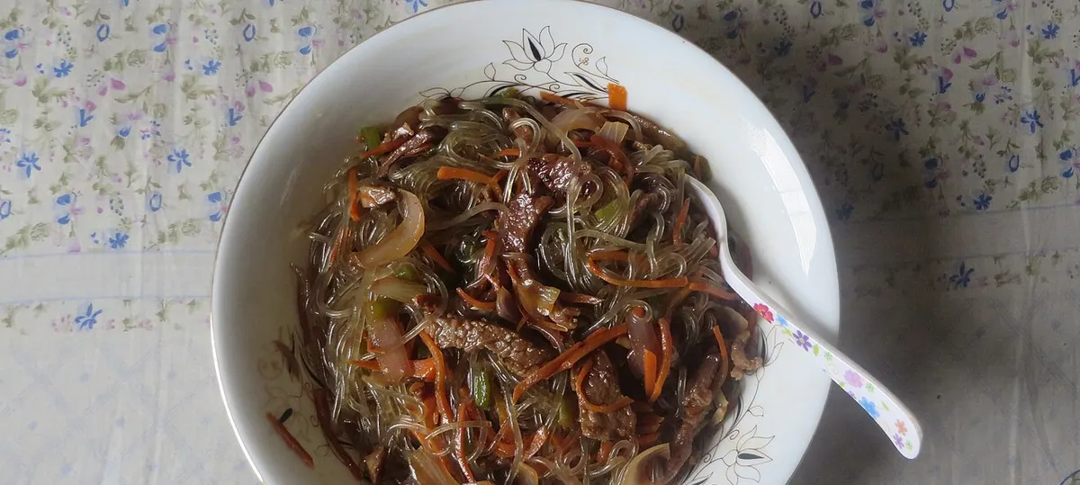 꽃무늬 접시에 담긴 집에서 만든 잡채. 당면과 소고기, 당근, 양파가 섞여 있고 숟가락이 걸쳐져 있는 모습