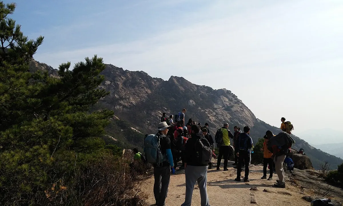 북한산 등산로 위에 배낭을 멘 등산객들이 모여 서 있고 뒤로 암봉 능선이 펼쳐진 모습