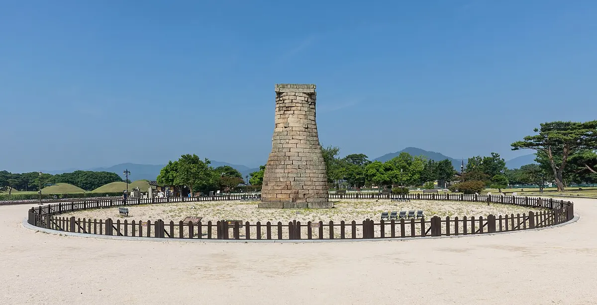 맑은 여름 하늘 아래 잔디밭 한가운데 서 있는 경주 첨성대와 그 주변을 두른 낮은 나무 울타리